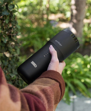 Electric Coffee Travel Mug juodas V2 - 355ml laikomas rankoje lauke su žaliuojančiu fonu.